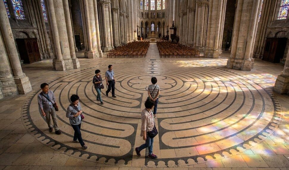シャルトル大聖堂の身廊にある床の迷宮（ラビリンス）を、ステンドグラスの光が差し込む中、アジア人観光客が静かに歩いている様子を捉えた写真。
