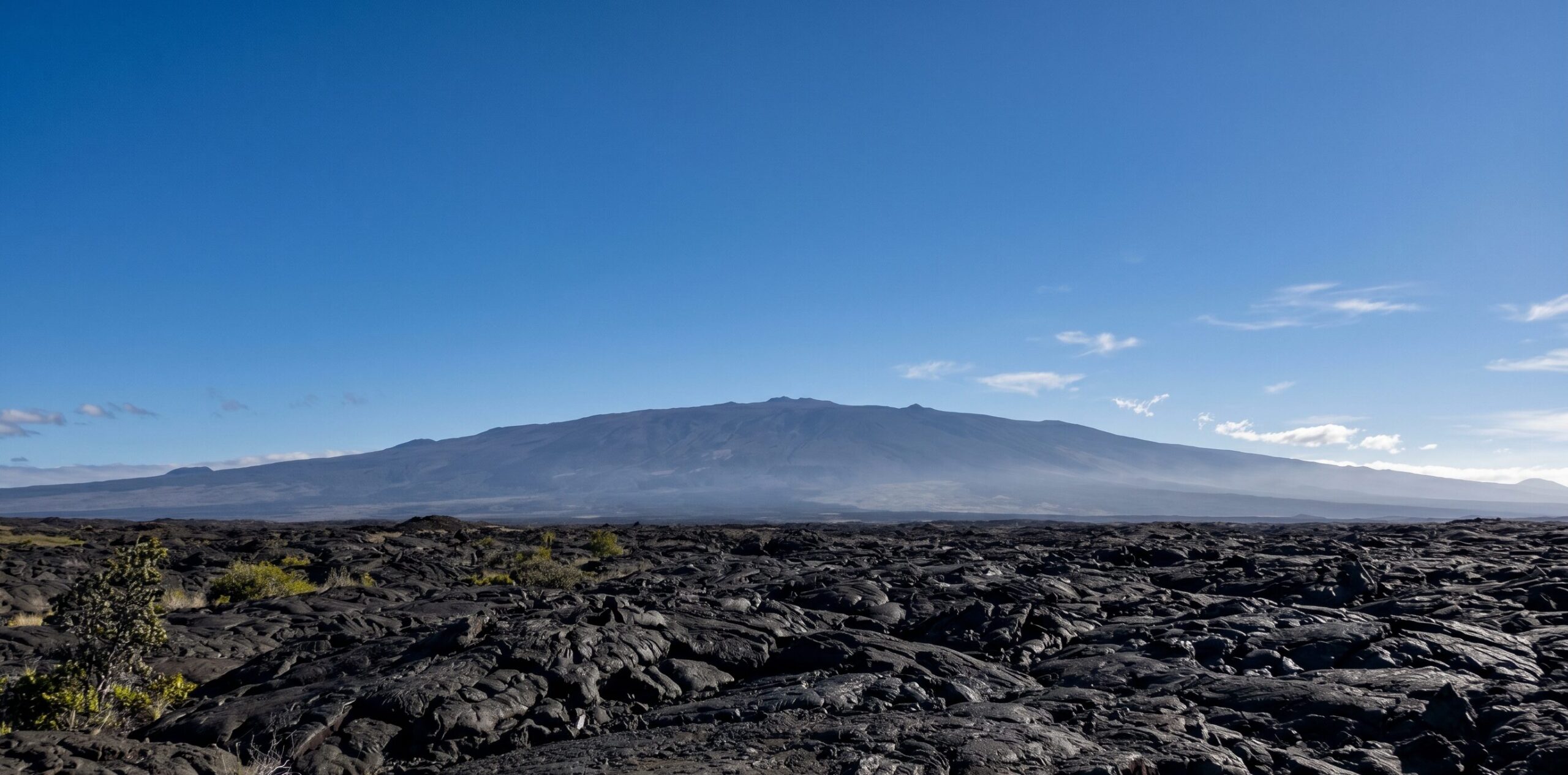 ハワイ島に広がる世界最大の火山マウナ・ロアの雄大な全景。黒い溶岩台地が地平線まで続き、盾状火山の巨大なスケール感が伝わる風景
